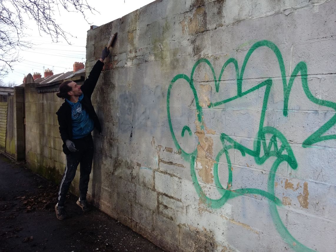 Muralist cleaning dirt and moss from a scruffy looking wall with a large tag on it