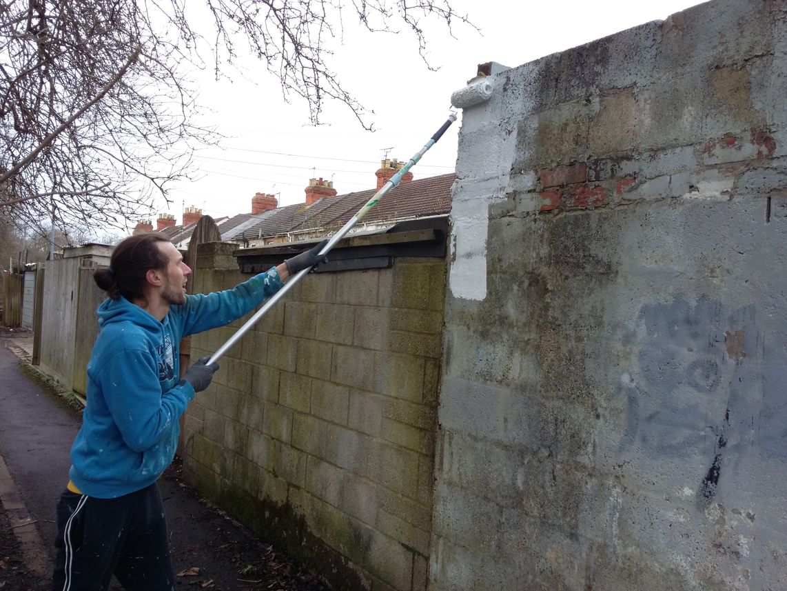 Muralist applying white paint to the wall with a roller on a pole