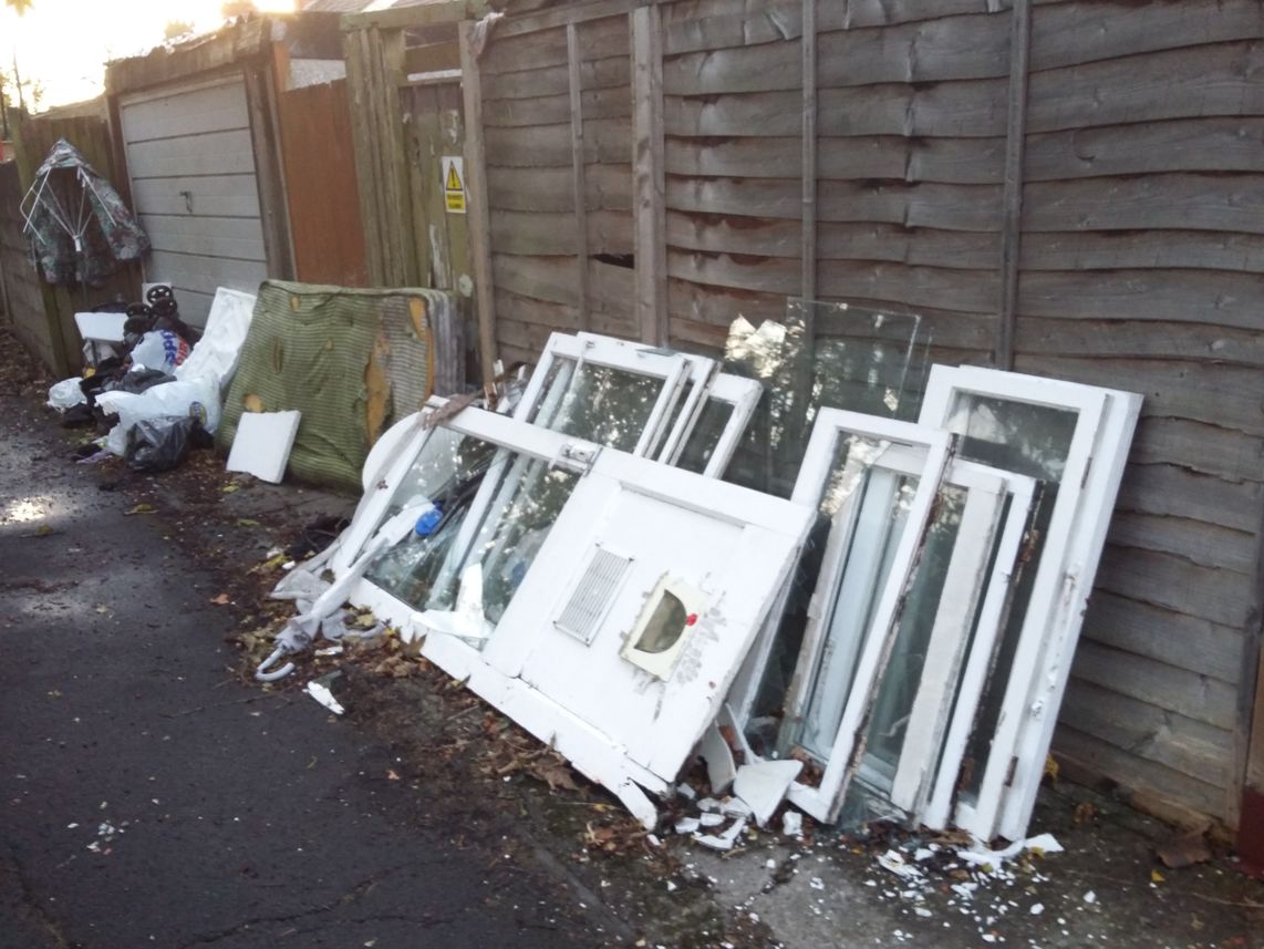 Flytipping in the alleyway at the back of a house.  Includes a door and windows.  Broken glass covers the ground.