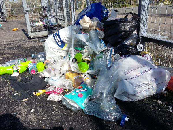 A pile of plastic bags and plastic water bottles by the metal fencing of a 5-a-side football pitch