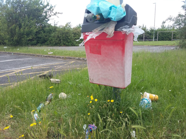 A small bin is overflowing.  Litter is in the long grass around it.