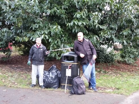 A litter picker playfully pokes another, who is leaning on a public bin, with their stick.  A bag of rubbish sits by the bin.