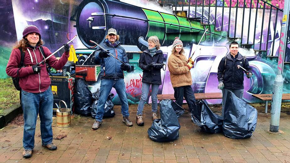A group of litter pickers in front of a graffiti mural of a train playfully wield their pickers at different angle next to their rubbish haul.  