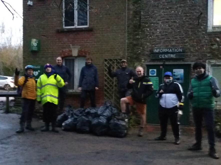 A group of 8 litter pickers stand waving by collected rubbish outside the Rangers Cottage at Coate Water