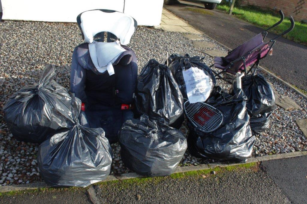 A pile of collected rubbish bags and loose items, including a tennis racket and pram, on a driveway