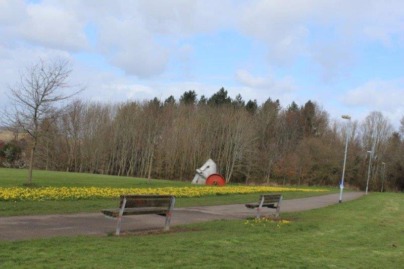 2 benches in front of a path.  An area of grass behind has flowers all along.  An area of woodland is in the background.