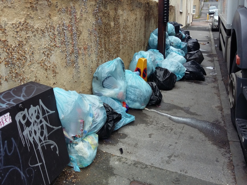 A large pile of rubbish bags on the pavement.  A lorry is parked in the road alongside.