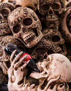 Many skulls together with ornate markings as of tribes people. There is a skeleton hand holding a bottle of Coca Cola, which is being tipped into the mouth of one of the skulls.