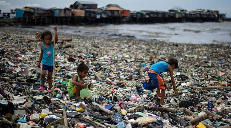 3 Asian children between around 6 and 8 years old traverse a beach covered in waste.  Huts / houses are distant in the background.