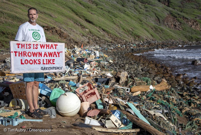 A man stands on an English beach covered in all sorts of litter, large and small.  He holds a placard that reads, 'This is what thrown away looks like - Greenpeace'