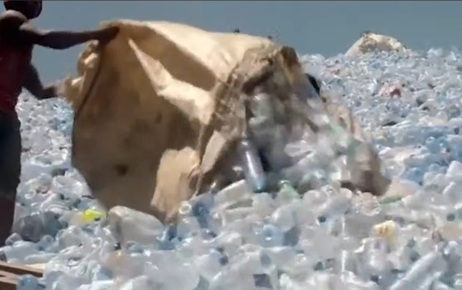 A man stands on a ginormous mountain of plastic bottles emptying a very large bag of plastic bottles onto it